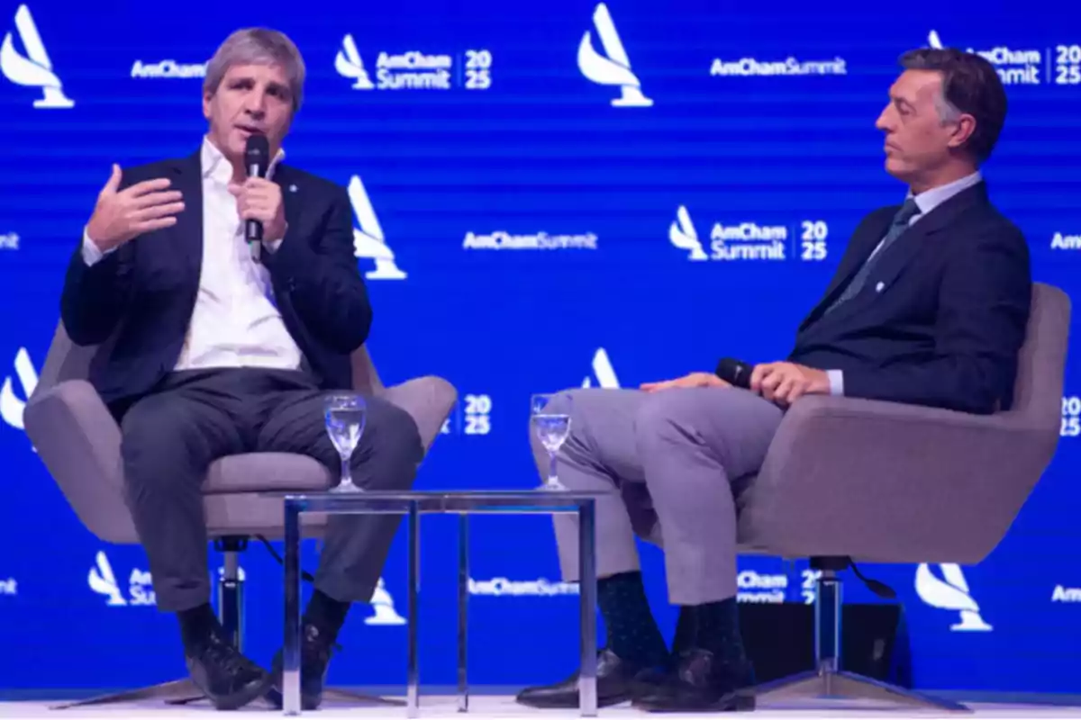Dos hombres sentados en sillones grises en un escenario con fondo azul durante un evento de AmCham Summit, uno de ellos sostiene un micrófono mientras habla.