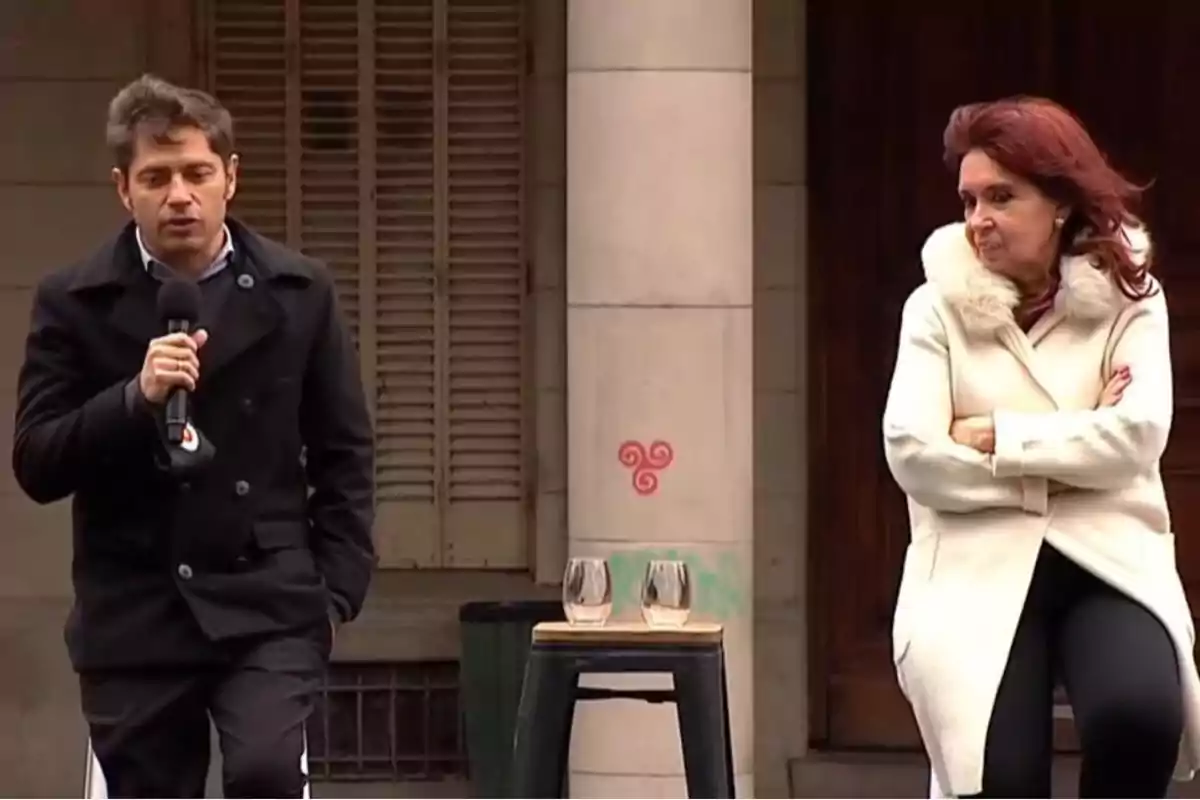 Dos personas están de pie al aire libre; una sostiene un micrófono y la otra tiene los brazos cruzados; hay una mesa con dos vasos frente a ellos.