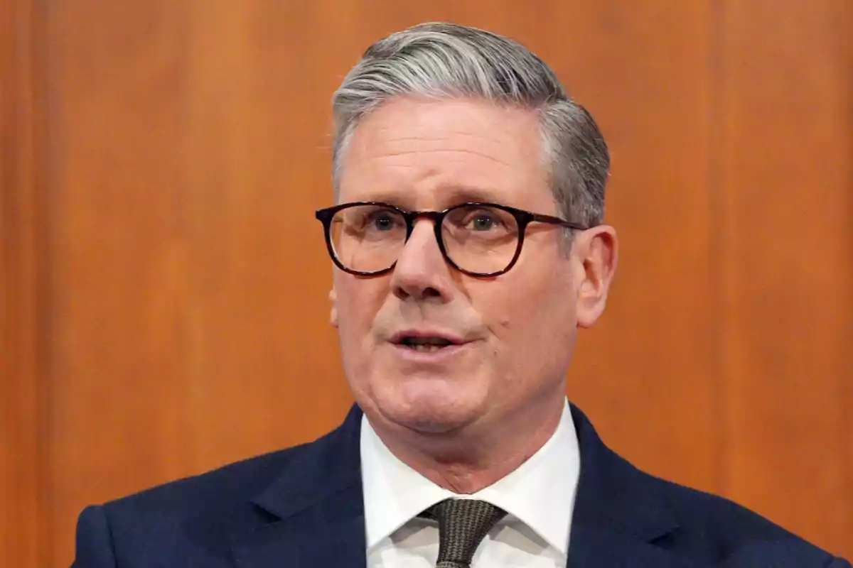 A man with glasses and gray hair is wearing a dark suit and tie in front of a wooden background. A man with glasses and gray hair is wearing a dark suit and tie in front of a wooden background.