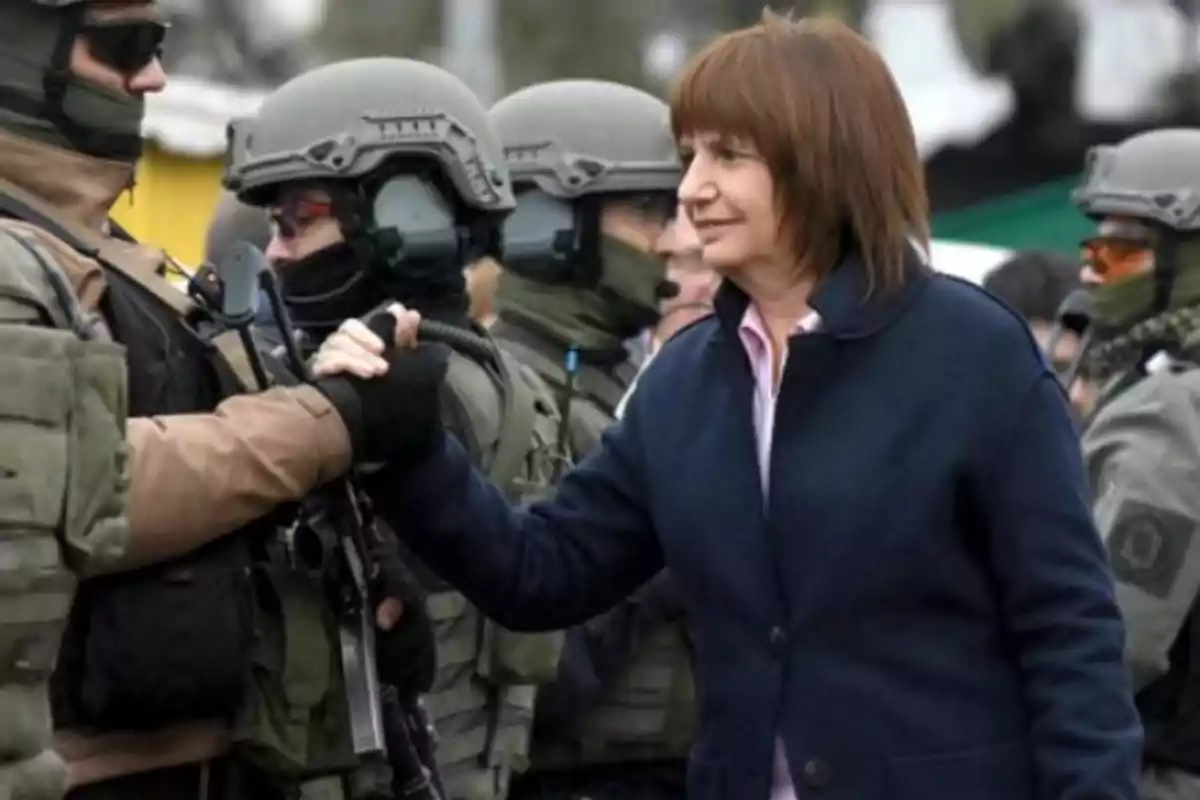 Una mujer estrecha la mano de un oficial de seguridad vestido con uniforme táctico y casco, mientras otros oficiales están alineados en el fondo. Una mujer estrecha la mano de un oficial de seguridad vestido con uniforme táctico y casco, mientras otros oficiales están alineados en el fondo.