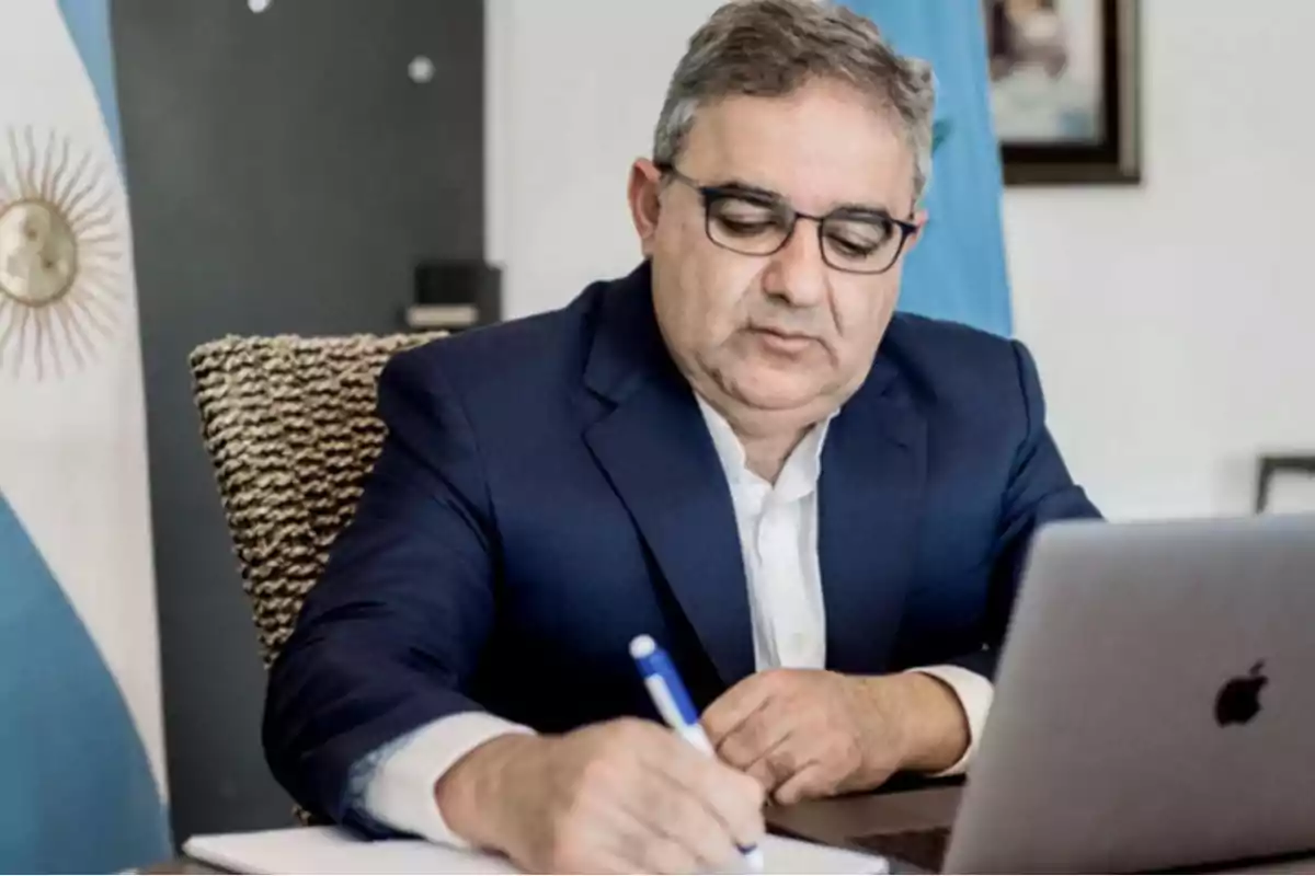 Hombre de traje azul escribiendo en un cuaderno mientras usa una computadora portátil.