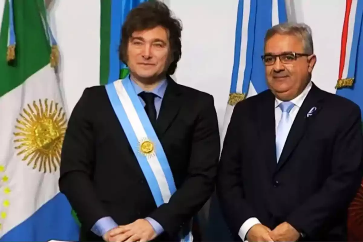 Dos hombres de pie con trajes oscuros y corbatas azules uno de ellos lleva una banda presidencial con banderas argentinas de fondo.
