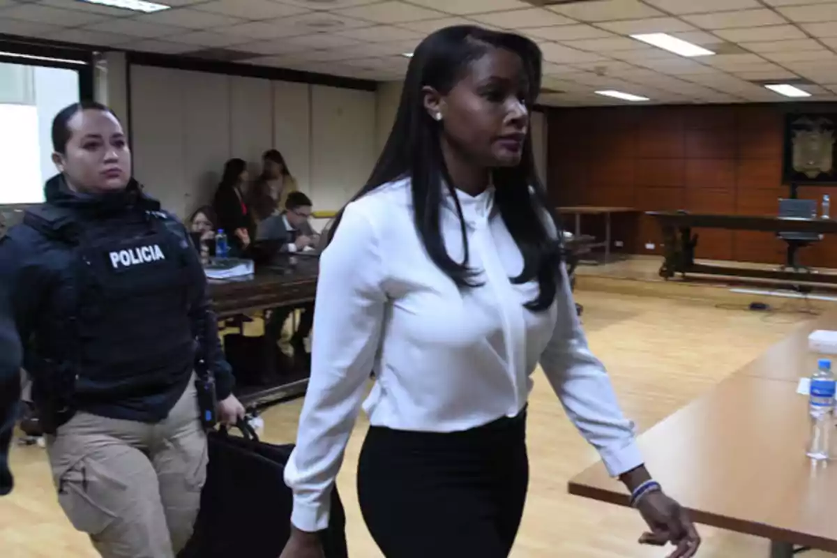 Una mujer vestida de blanco camina junto a una oficial de policía en una sala de audiencias.