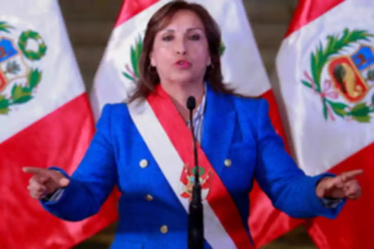 Una persona con un traje azul y una banda presidencial habla frente a micrófonos con banderas de Perú en el fondo. Una persona con un traje azul y una banda presidencial habla frente a micrófonos con banderas de Perú en el fondo.