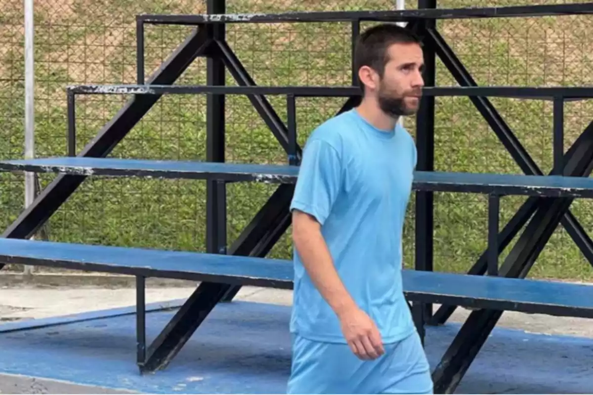 Un hombre con camiseta y pantalones cortos azules camina frente a unas gradas metálicas en un área al aire libre. Un hombre con camiseta y pantalones cortos azules camina frente a unas gradas metálicas en un área al aire libre.