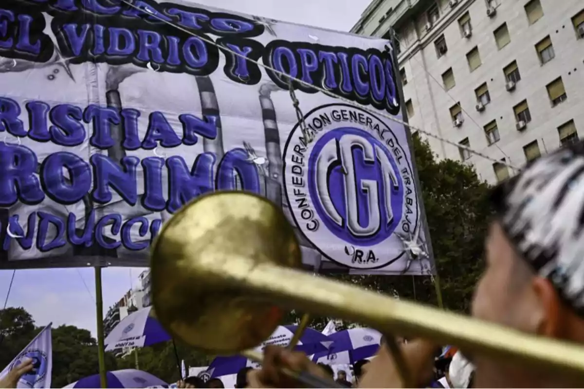 Una persona toca un instrumento de metal frente a una pancarta de la Confederación General del Trabajo durante una manifestación en una ciudad con edificios de fondo.
