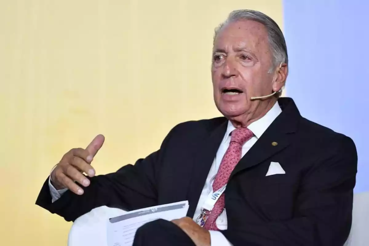 Hombre mayor con traje oscuro y corbata rosa hablando en un evento con micrófono de diadema. Hombre mayor con traje oscuro y corbata rosa hablando en un evento con micrófono de diadema.