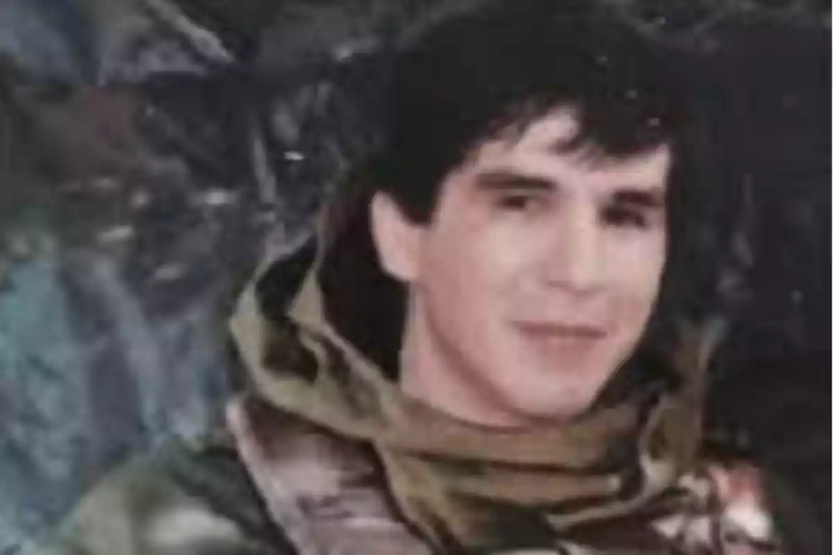 Un hombre joven con cabello oscuro lleva una chaqueta de camuflaje y está sonriendo ligeramente frente a un fondo oscuro. Un hombre joven con cabello oscuro lleva una chaqueta de camuflaje y está sonriendo ligeramente frente a un fondo oscuro.