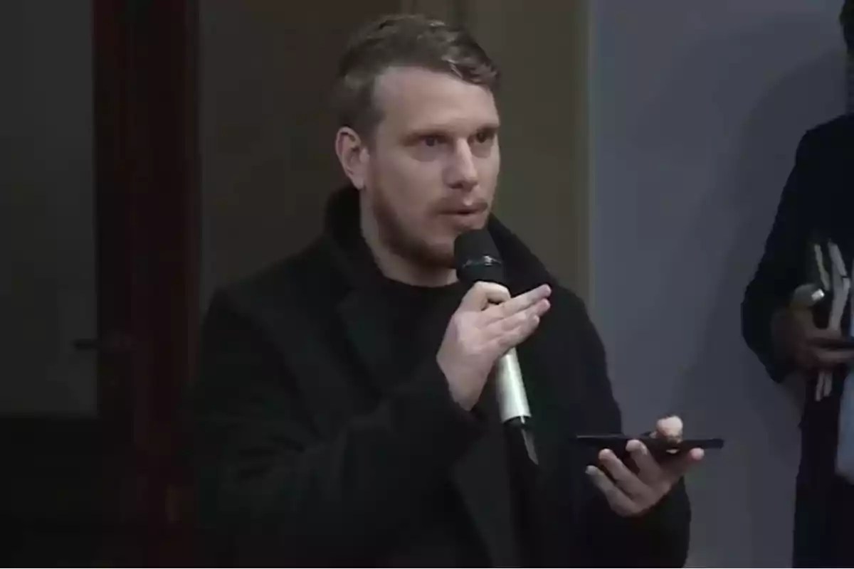 Un hombre con barba y cabello corto sostiene un micrófono mientras parece estar hablando en un evento. Un hombre con barba y cabello corto sostiene un micrófono mientras parece estar hablando en un evento.