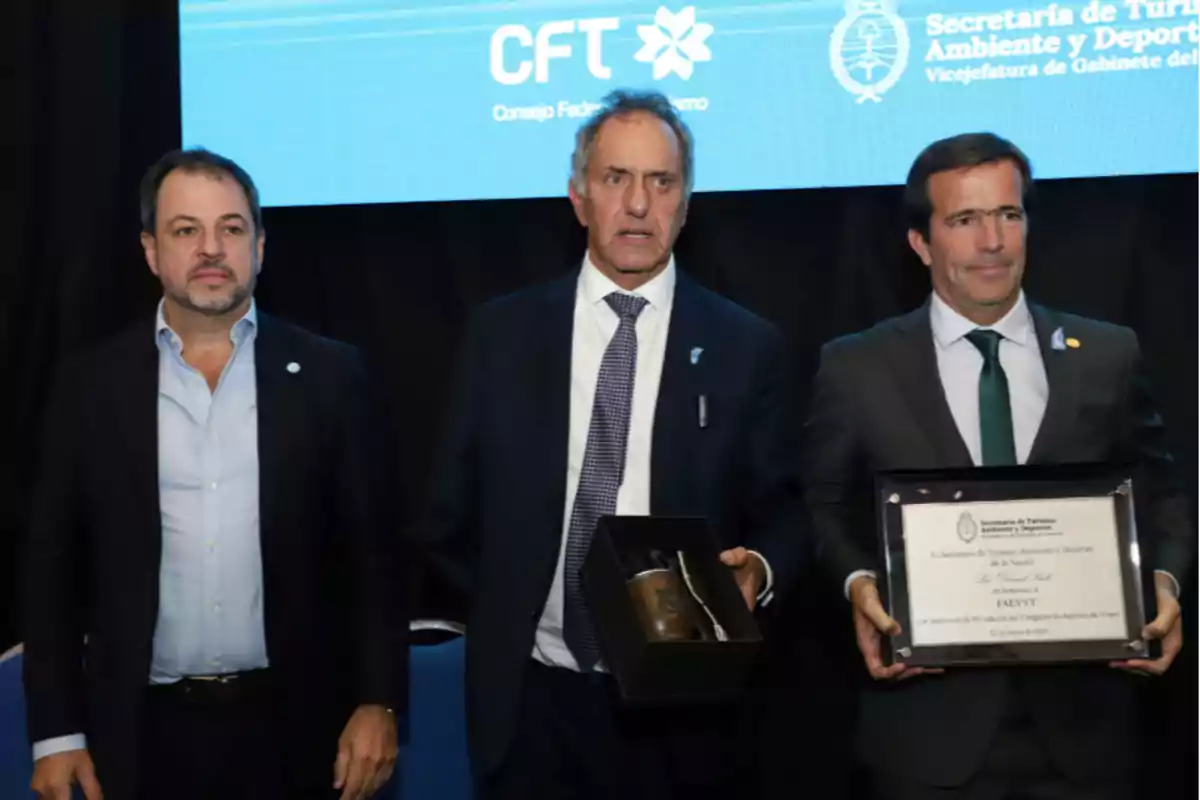 Tres hombres de traje posan en un evento, uno de ellos sostiene un mate en una caja y otro una placa conmemorativa, con un fondo que muestra logotipos de una organización relacionada con turismo. Tres hombres de traje posan en un evento, uno de ellos sostiene un mate en una caja y otro una placa conmemorativa, con un fondo que muestra logotipos de una organización relacionada con turismo.