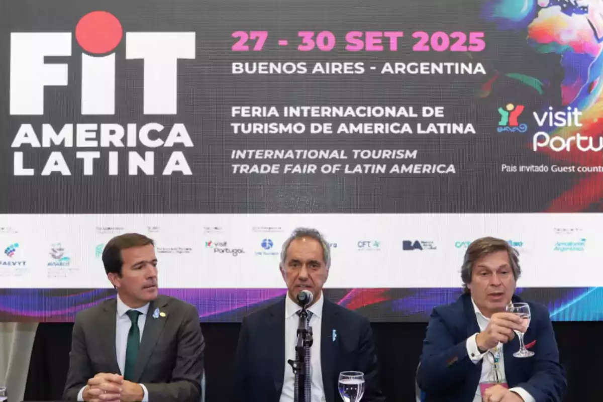 Tres hombres sentados en una mesa durante una conferencia de prensa, con un cartel detrás que anuncia la Feria Internacional de Turismo de América Latina en Buenos Aires del 27 al 30 de septiembre de 2025, destacando a Portugal como país invitado.