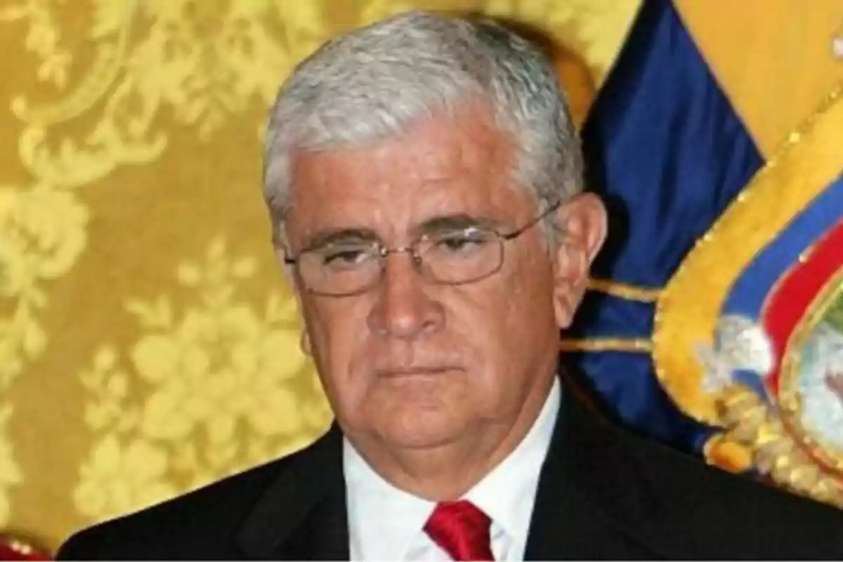 Un hombre de cabello canoso y gafas con expresión seria, vestido con traje oscuro y corbata roja, frente a un fondo decorativo dorado y azul. Un hombre de cabello canoso y gafas con expresión seria, vestido con traje oscuro y corbata roja, frente a un fondo decorativo dorado y azul.