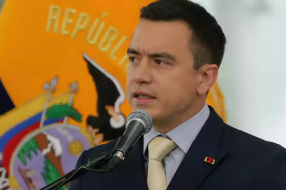A man speaking in front of a microphone with a background showing part of a shield and the word A man speaking in front of a microphone with a background showing part of a shield and the word