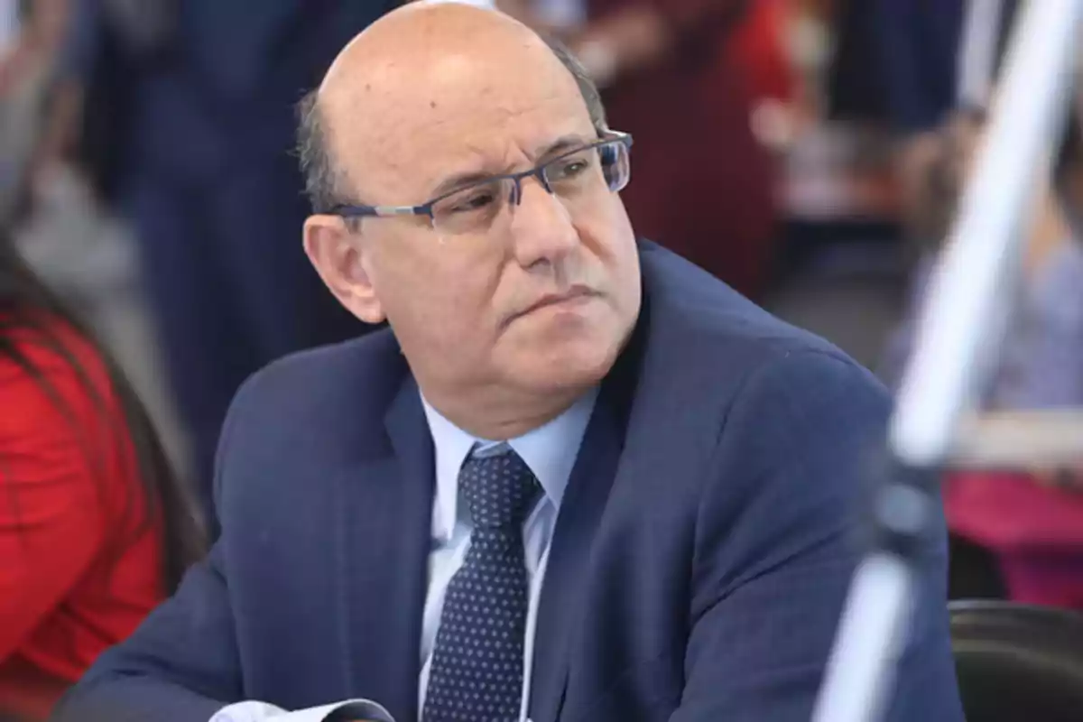 A man with glasses and a blue suit looks to the side in a professional setting.