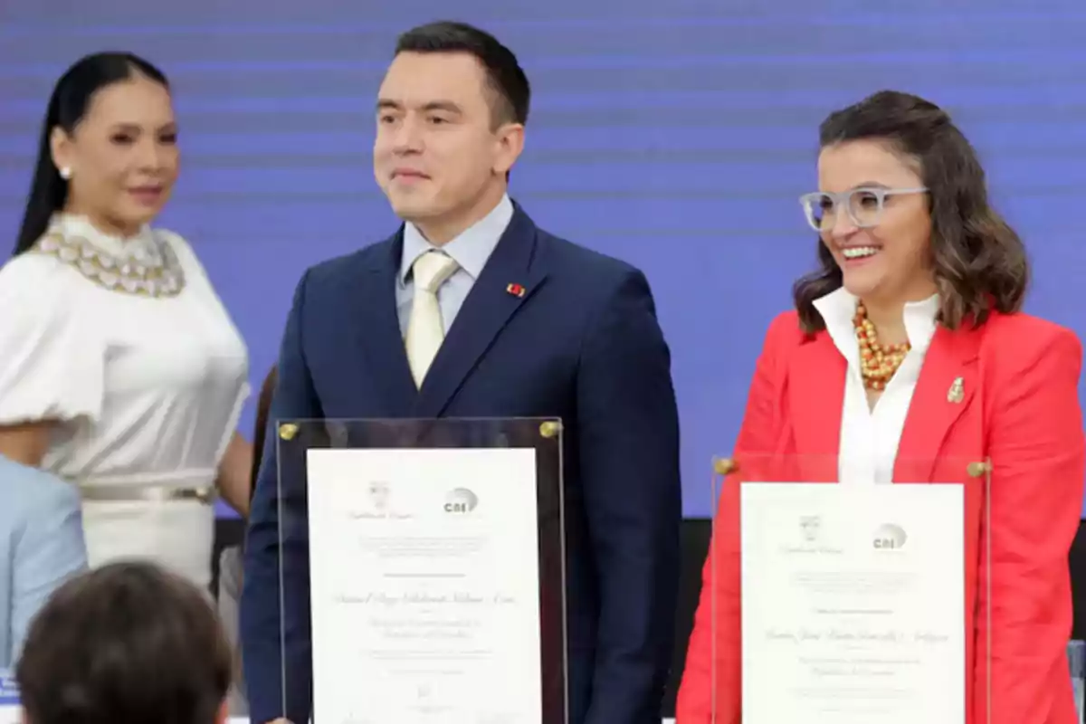 Two people hold diplomas at a formal event while a woman watches in the background.