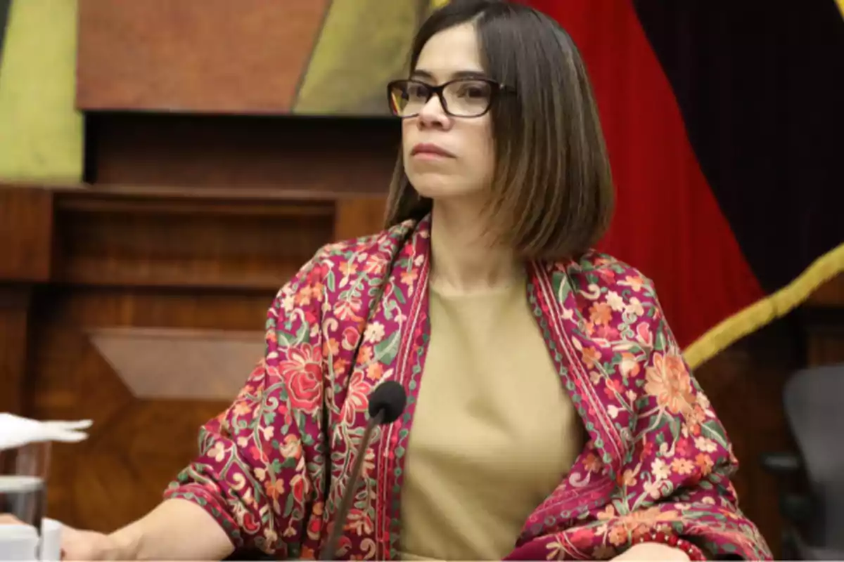 A person with glasses and short hair is wearing a colorful shawl while sitting in a room with a microphone in front of her.