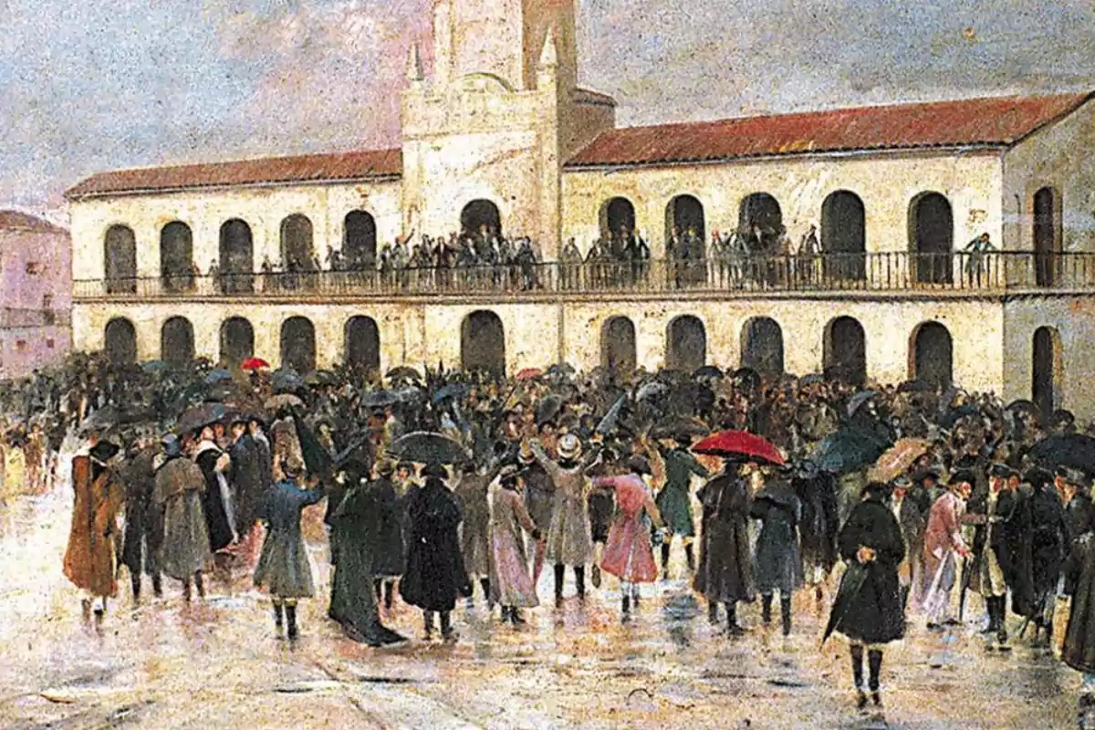 A crowd gathers in front of a colonial building with balconies while some people hold colorful umbrellas.