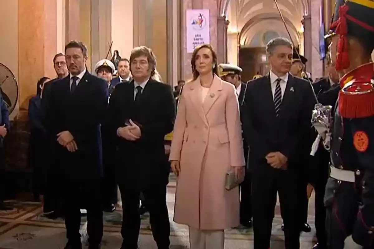Un grupo de personas vestidas formalmente se encuentra en un evento oficial en un edificio con arquitectura clásica, mientras un guardia con uniforme ceremonial está presente.