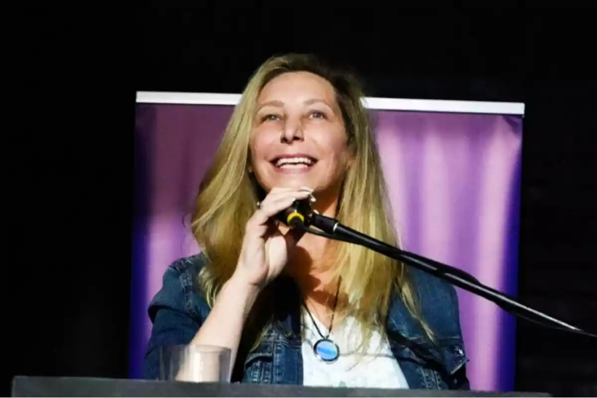 Smiling blonde woman speaking into a microphone with a dark purple background