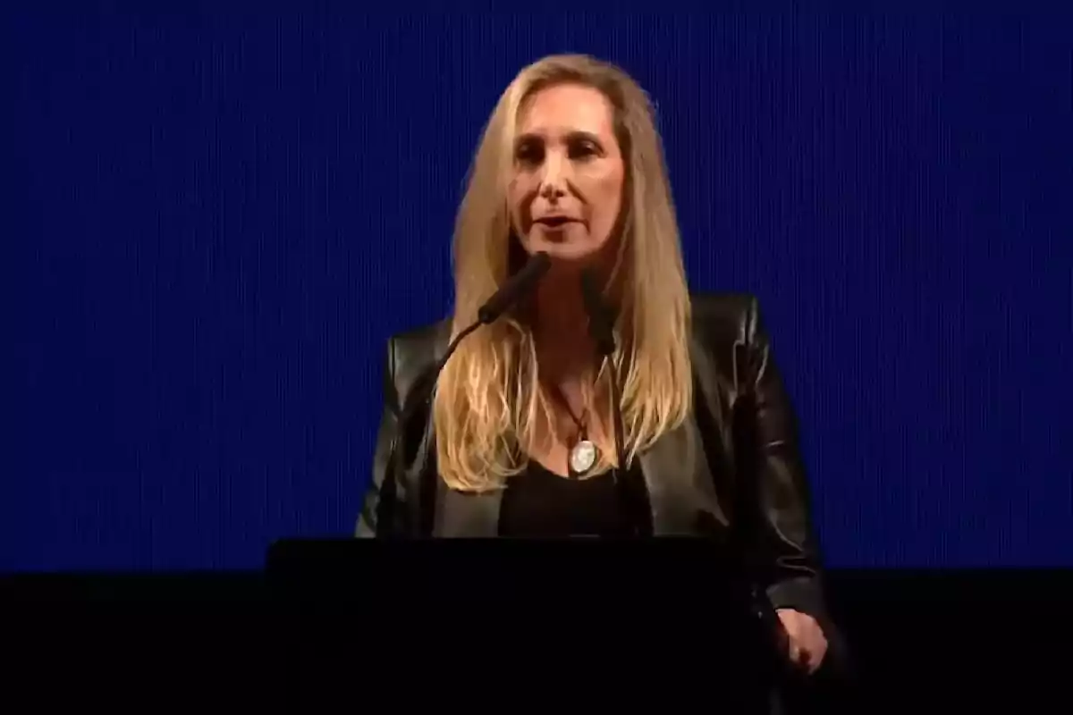 Mujer rubia con chaqueta negra hablando frente a un micrófono con fondo azul Mujer rubia con chaqueta negra hablando frente a un micrófono con fondo azul