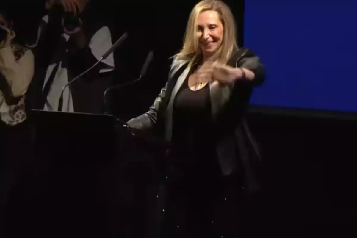 Mujer rubia sonriente en un escenario vestida de negro y chaqueta oscura frente a un podio con micrófonos Mujer rubia sonriente en un escenario vestida de negro y chaqueta oscura frente a un podio con micrófonos