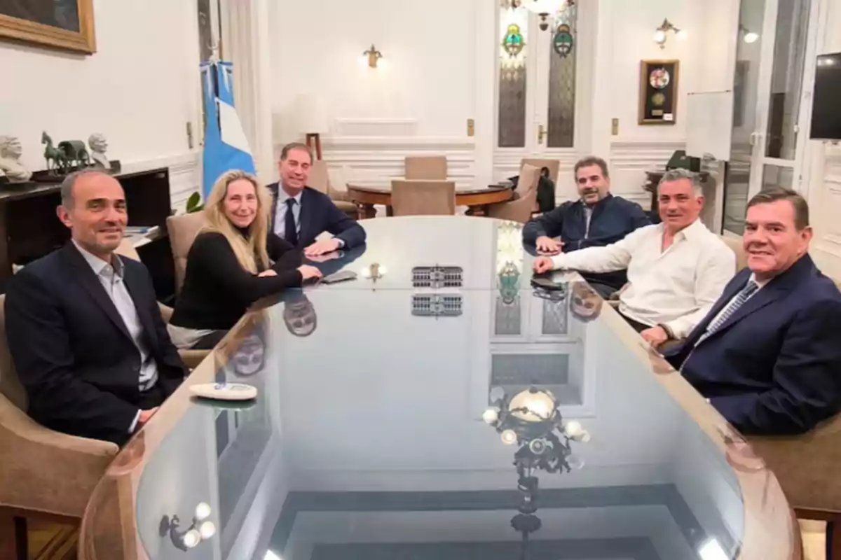 Siete personas sentadas alrededor de una mesa de vidrio en una sala elegante con una bandera argentina al fondo