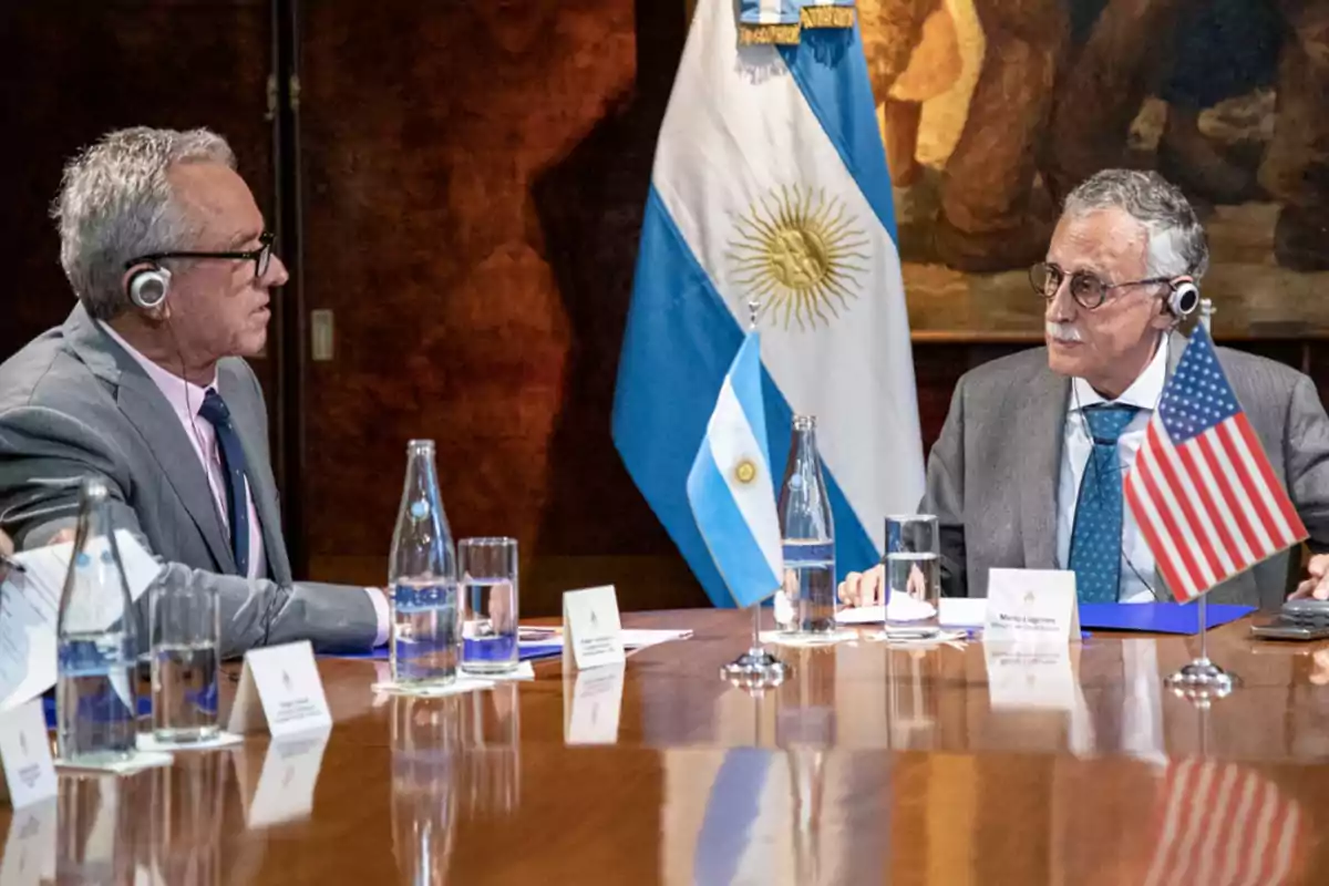 Two men with translation headsets are talking at a conference table with Argentine and United States flags, water bottles, and papers on the table.
