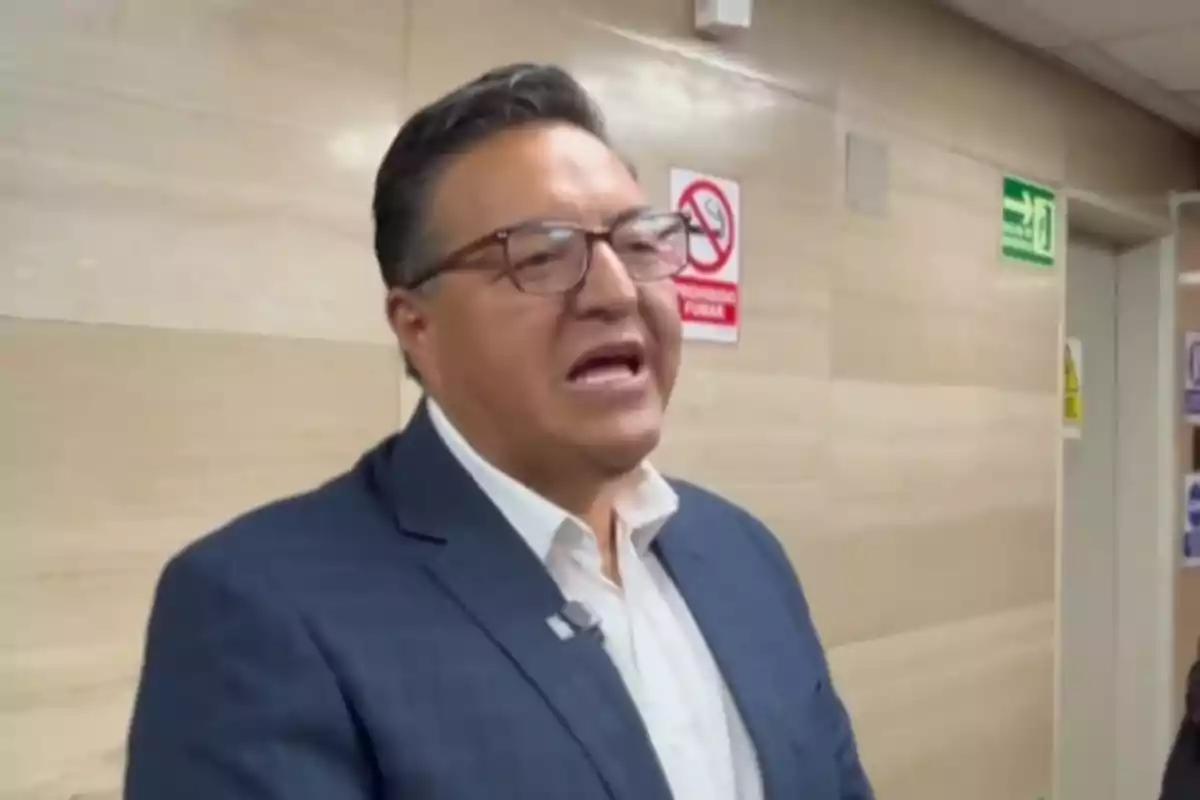 Man with glasses and a blue blazer talking in a building hallway with signs on the wall Man with glasses and a blue blazer talking in a building hallway with signs on the wall