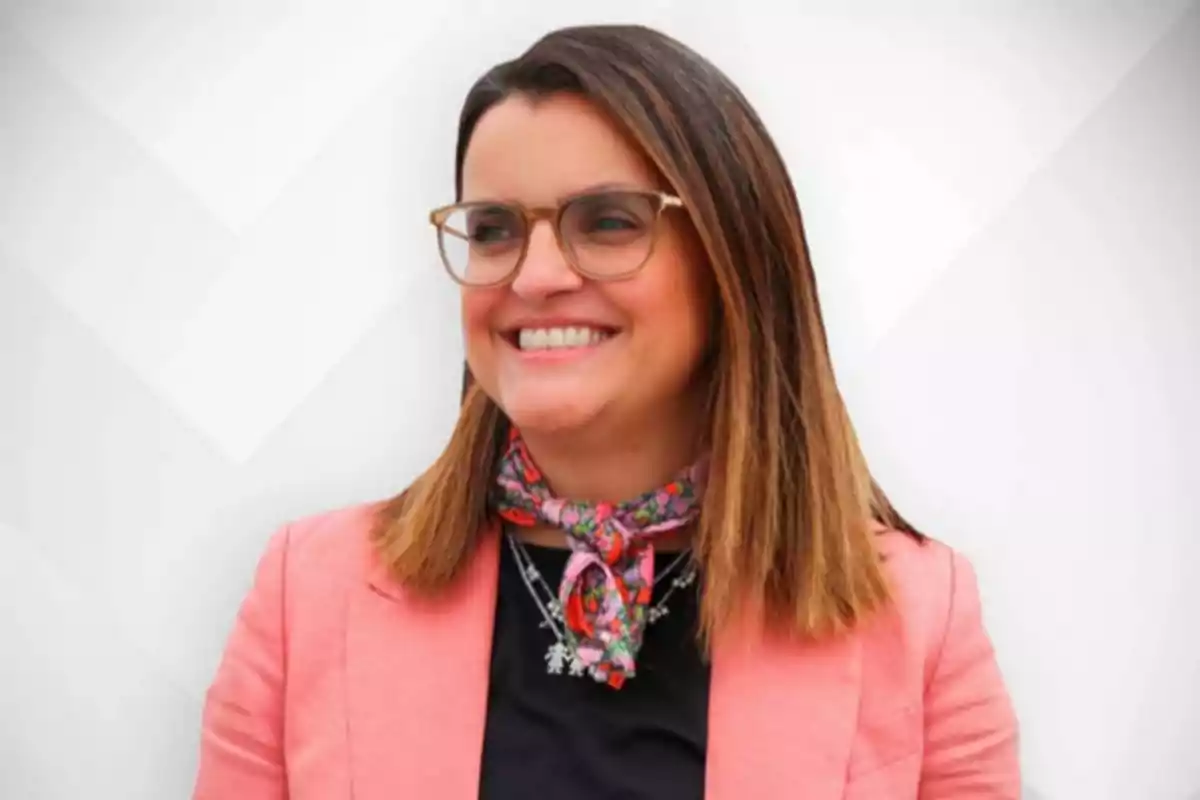 Mujer sonriente con gafas y cabello lacio, lleva un saco rosa, blusa negra y un pañuelo colorido en el cuello, fondo claro geométrico Mujer sonriente con gafas y cabello lacio, lleva un saco rosa, blusa negra y un pañuelo colorido en el cuello, fondo claro geométrico