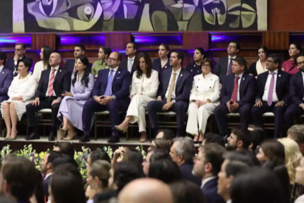 Group of people dressed formally seated in an auditorium during an official event with an abstract mural on the back wall