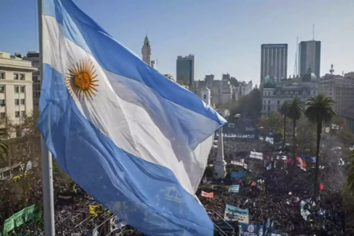 Argentine flag waving in front of a crowd gathered in a downtown Buenos Aires square, with buildings and palm trees in the background Argentine flag waving in front of a crowd gathered in a downtown Buenos Aires square, with buildings and palm trees in the background