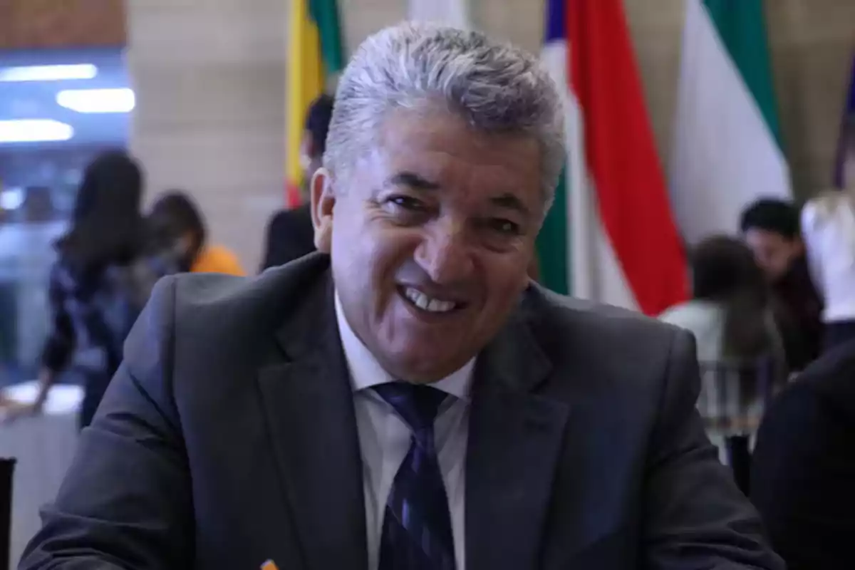 Gray-haired man in a dark suit smiling at an event with flags in the background