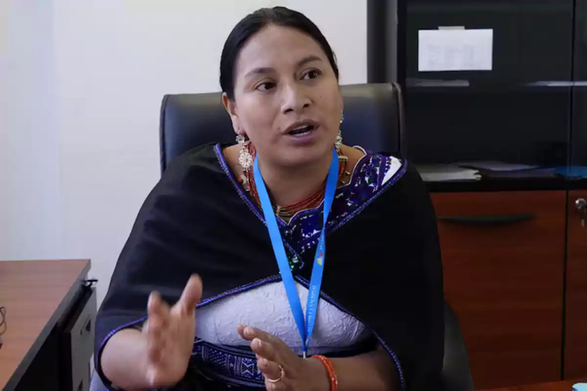 Indigenous woman sitting in an office, talking and gesturing with her hands, wearing traditional clothing and a blue badge.