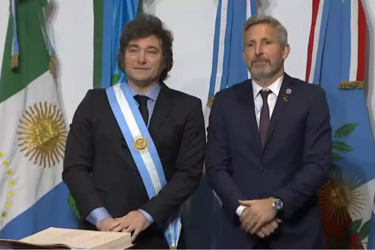 Dos hombres de traje posan frente a varias banderas argentinas uno de ellos lleva la banda presidencial y una medalla dorada
