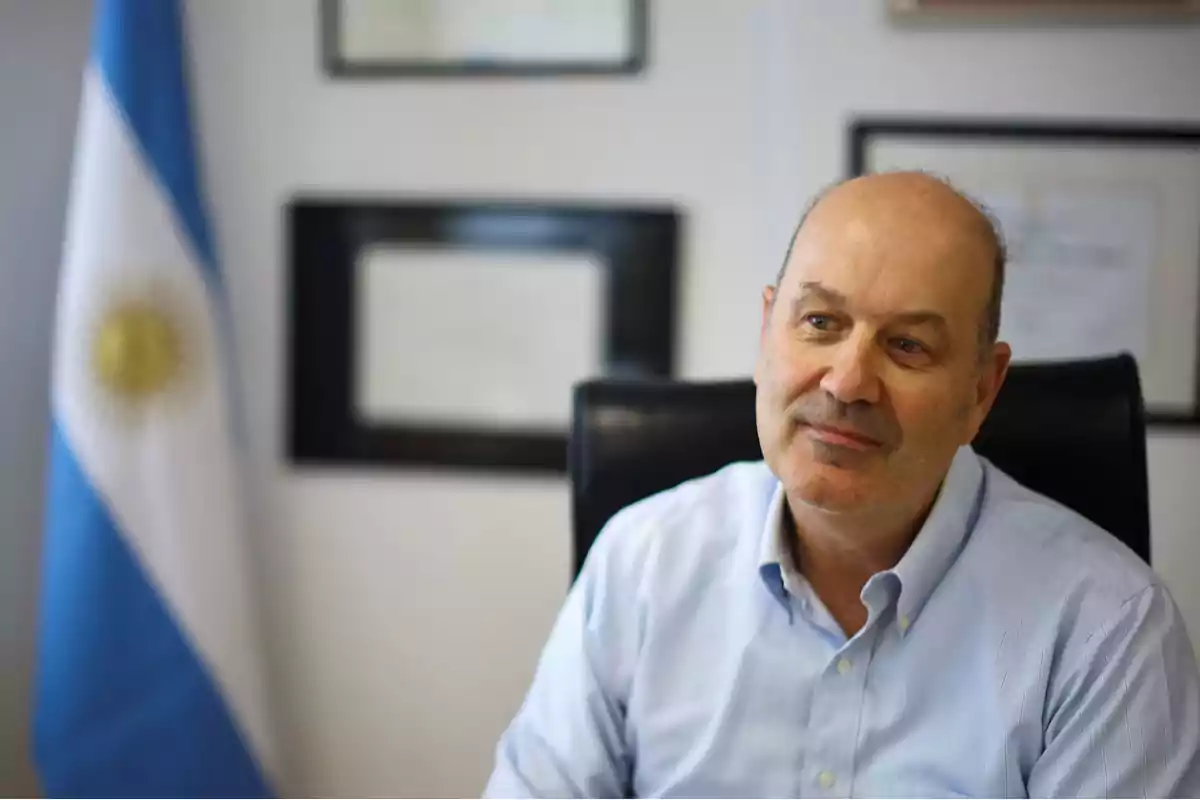 Hombre calvo de mediana edad con camisa celeste sentado en una oficina con una bandera de Argentina y diplomas en la pared