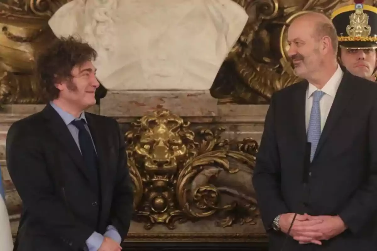 Dos hombres de traje se miran y sonríen en un entorno elegante con decoración dorada y una escultura blanca al fondo Dos hombres de traje se miran y sonríen en un entorno elegante con decoración dorada y una escultura blanca al fondo