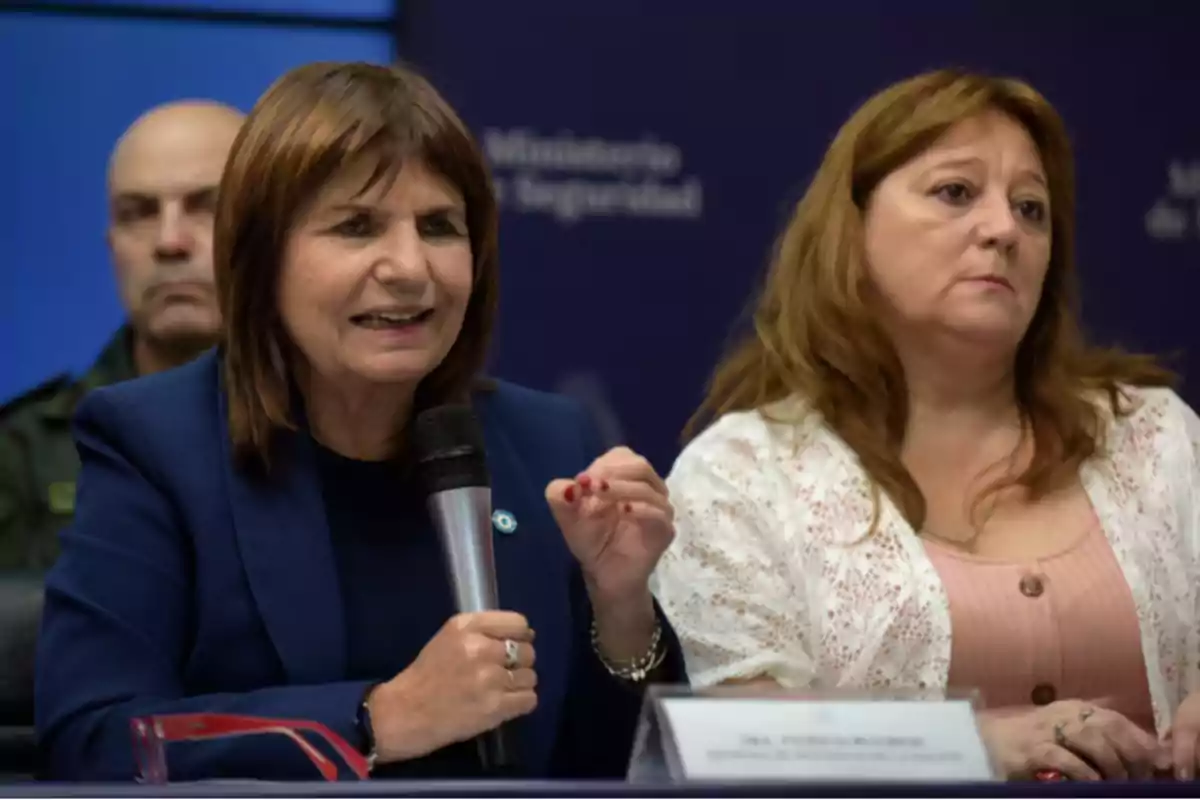 Mujer de cabello castaño hablando en una conferencia de prensa mientras sostiene un micrófono, acompañada por otra mujer y un hombre al fondo Mujer de cabello castaño hablando en una conferencia de prensa mientras sostiene un micrófono, acompañada por otra mujer y un hombre al fondo