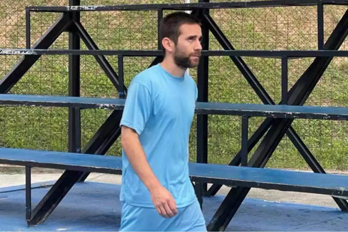 Hombre con camiseta y pantalón azul claro caminando frente a gradas metálicas en una cancha al aire libre Hombre con camiseta y pantalón azul claro caminando frente a gradas metálicas en una cancha al aire libre