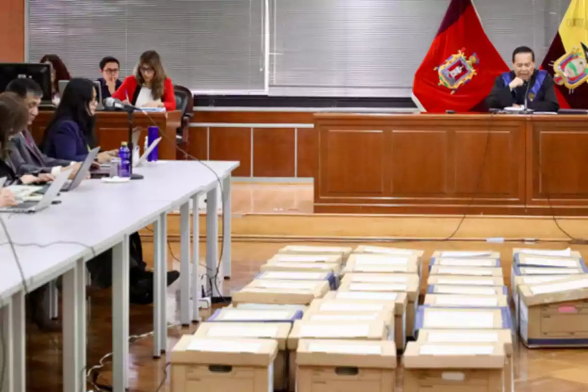Sala de audiencias con varias personas trabajando en computadoras y numerosas cajas de documentos en el suelo frente a un juez