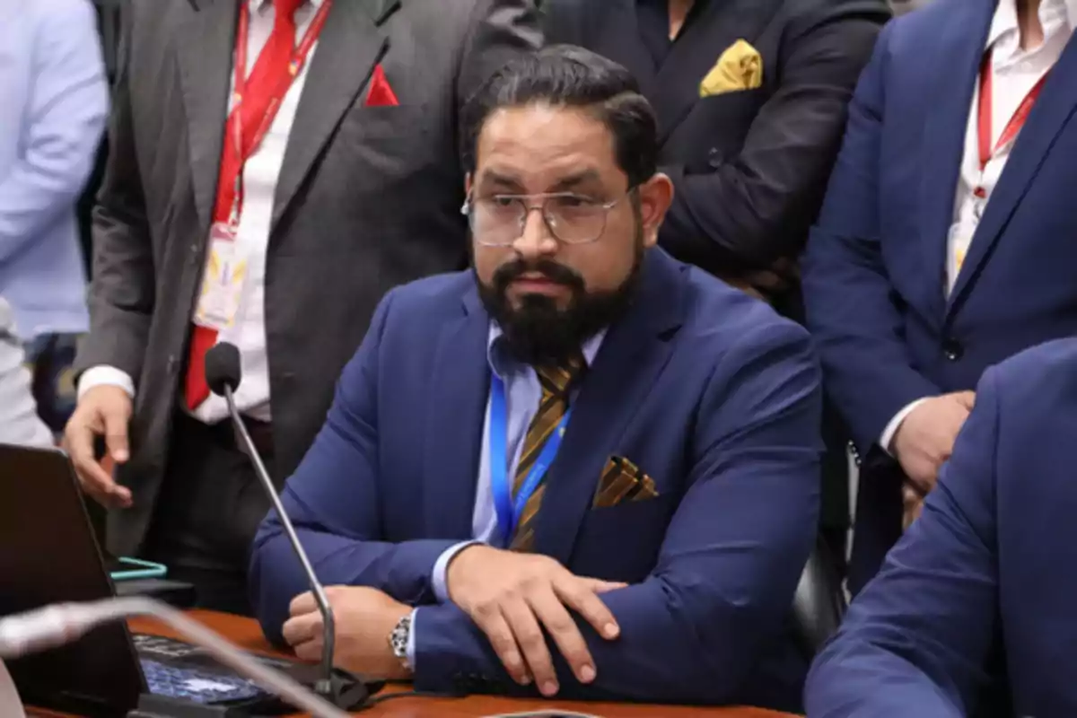Hombre con barba y gafas usando traje azul marino y corbata dorada sentado frente a un micrófono en una reunión formal rodeado de otras personas con vestimenta de negocios