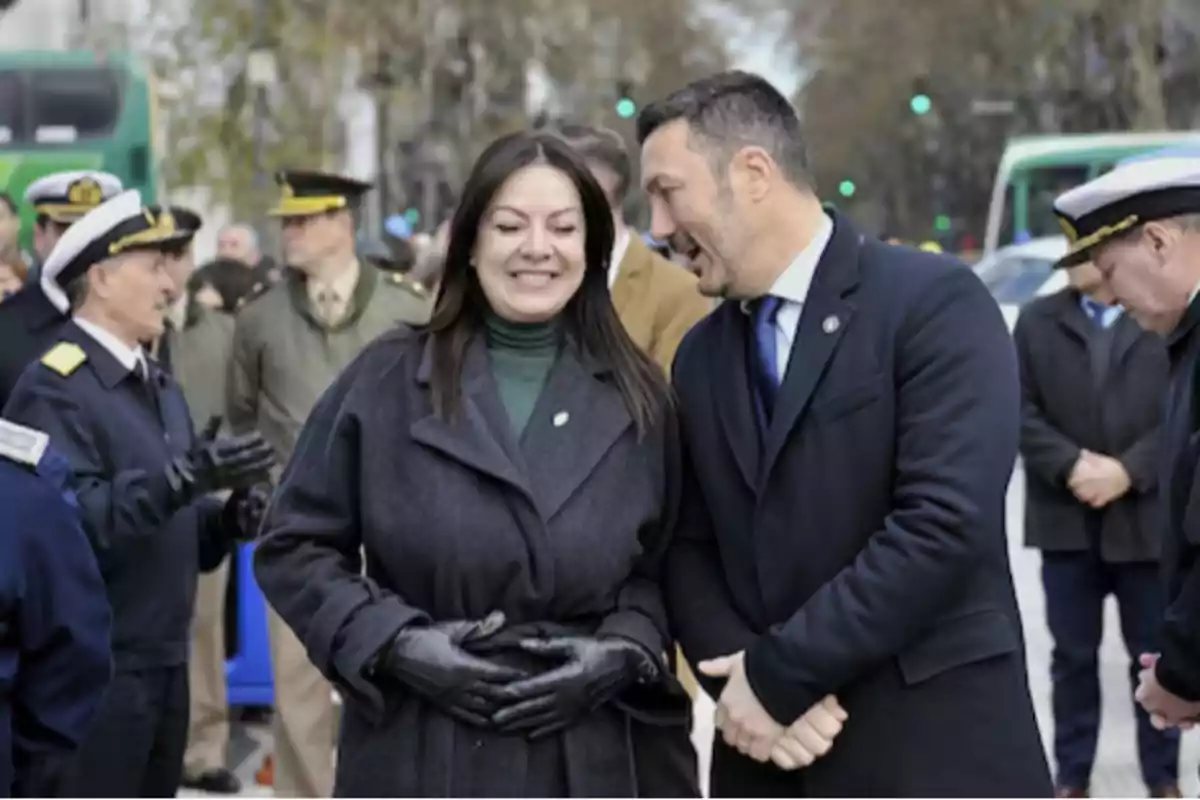 Dos personas conversan y sonríen en un evento al aire libre rodeados de oficiales con uniforme Dos personas conversan y sonríen en un evento al aire libre rodeados de oficiales con uniforme