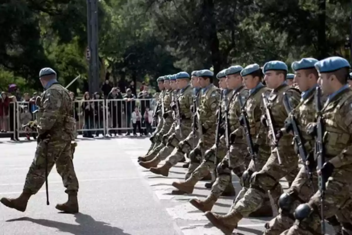 Soldados marchando en formación con boinas celestes y uniformes de camuflaje durante un desfile frente a espectadores detrás de una valla