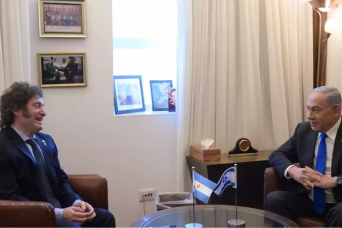 Two men in suits are talking while seated in an elegant office with light curtains and the flags of Argentina and Israel on a round table. Two men in suits are talking while seated in an elegant office with light curtains and the flags of Argentina and Israel on a round table.