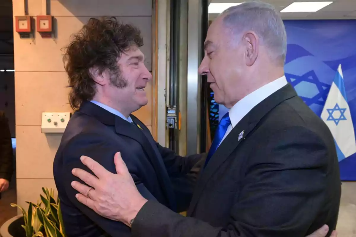Two men in suits greet each other warmly in front of an Israeli flag in an indoor setting. Two men in suits greet each other warmly in front of an Israeli flag in an indoor setting.