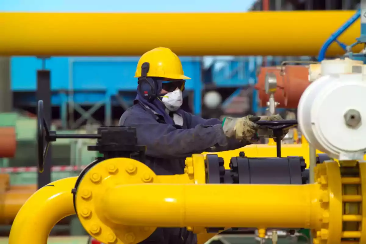 Industrial worker with a helmet and mask operating a valve at a gas plant with yellow pipes