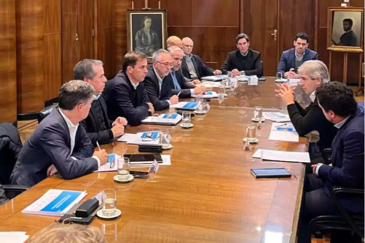 A group of people in suits sitting around a wooden table in a meeting room, with documents, cups, and glasses of water on the table and two portraits hanging on the back wall.