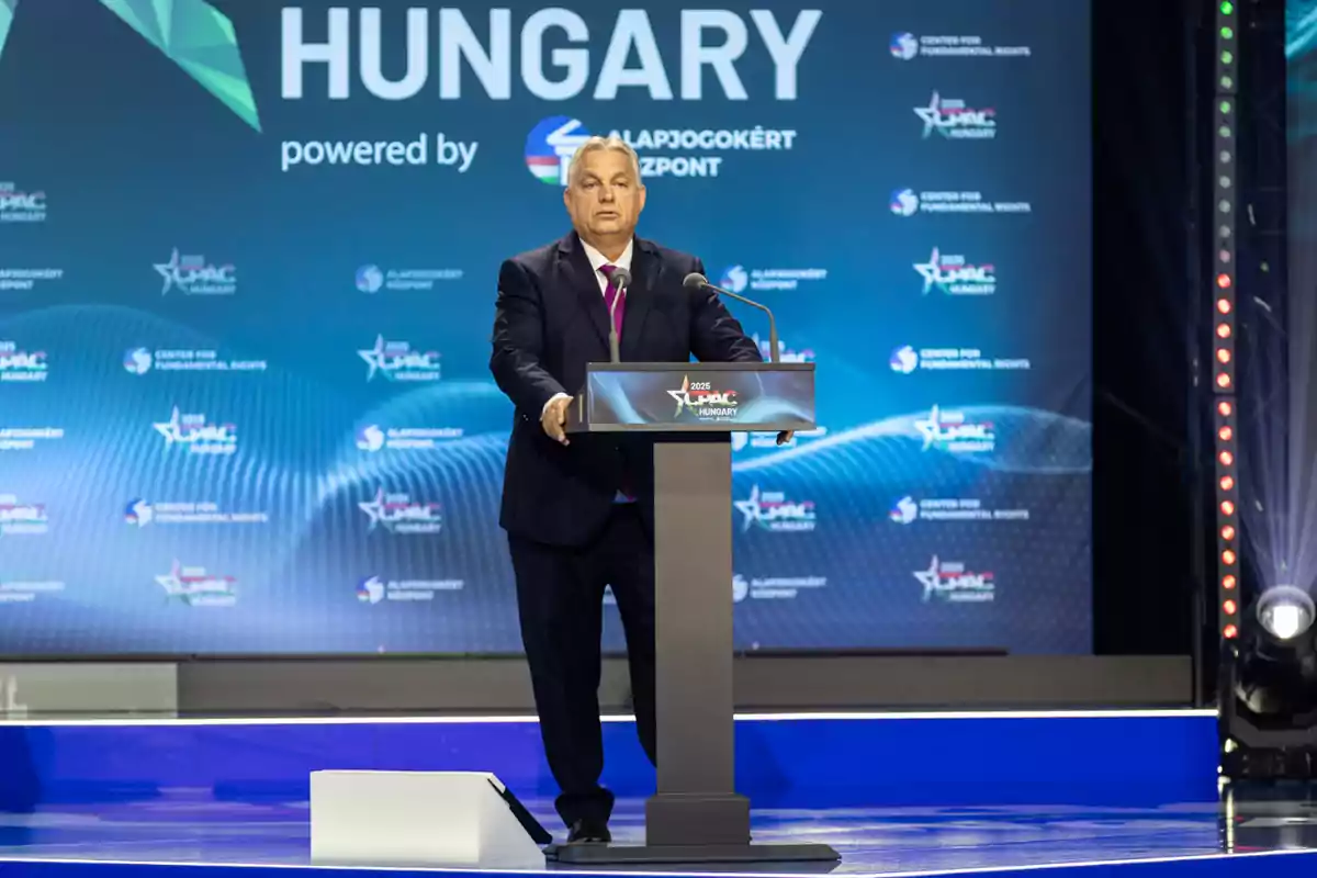 A man in a dark suit speaks at a podium during an event in Hungary with a blue background displaying logos and the name of the country.