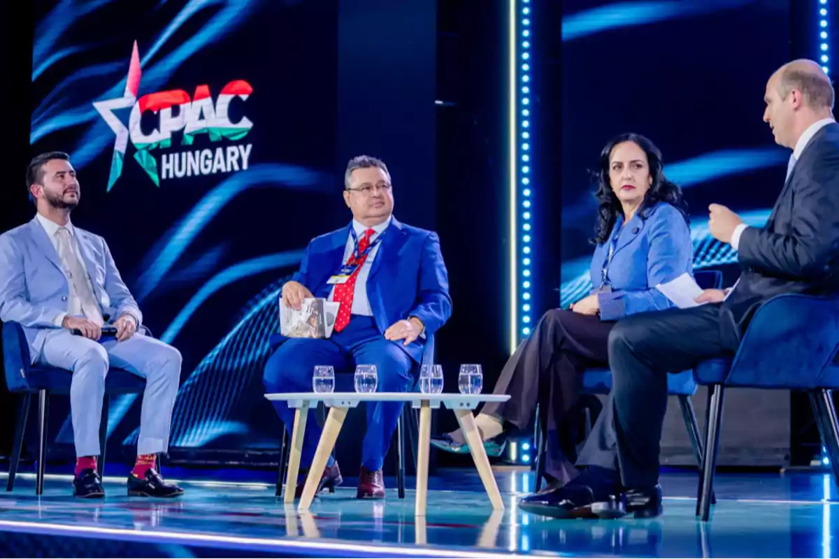 Four people in formal attire are seated on a discussion panel on a stage with the CPAC Hungary logo in the background and a low table with glasses of water in front of them. Four people in formal attire are seated on a discussion panel on a stage with the CPAC Hungary logo in the background and a low table with glasses of water in front of them.