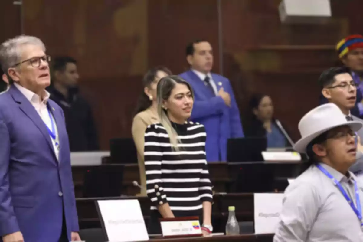 Varias personas de pie en un recinto parlamentario, algunas con credenciales y una mujer en el centro con un suéter de rayas blancas y negras