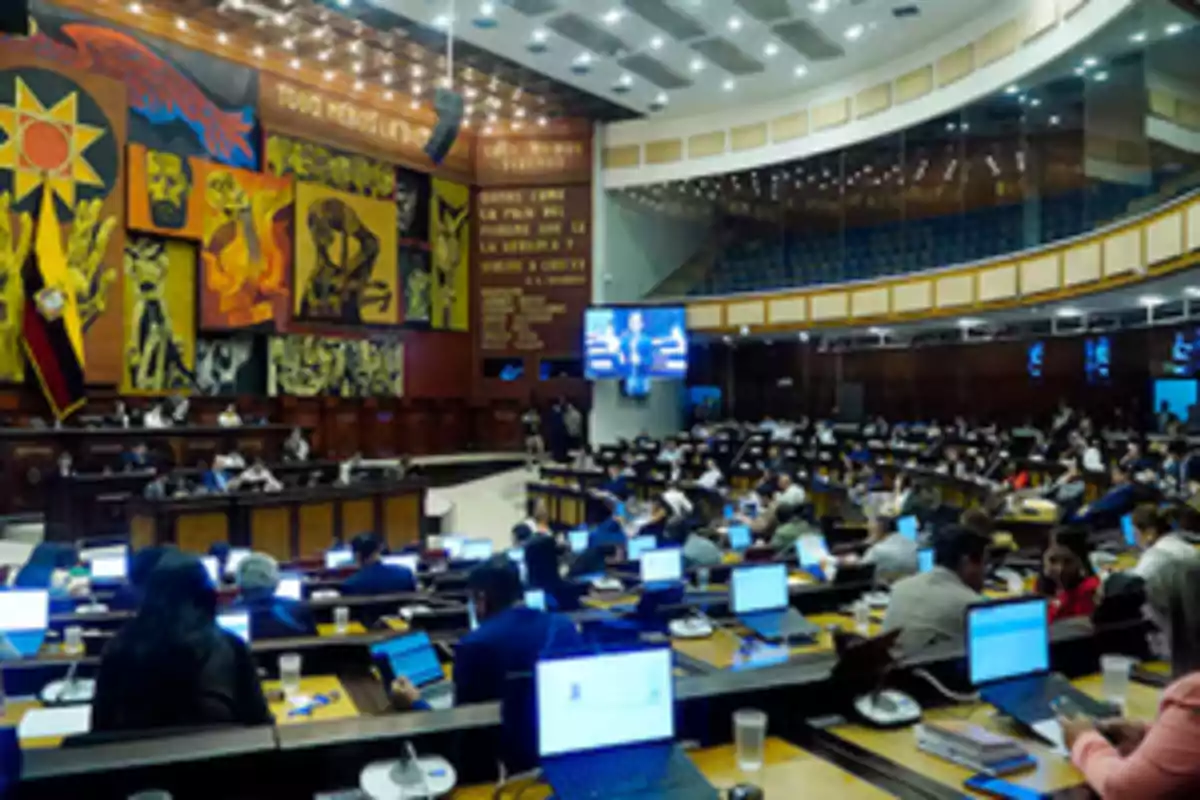 Vista general del pleno de la Asamblea Nacional de Ecuador con legisladores trabajando en sus escritorios y computadoras bajo un mural colorido y la bandera nacional Vista general del pleno de la Asamblea Nacional de Ecuador con legisladores trabajando en sus escritorios y computadoras bajo un mural colorido y la bandera nacional